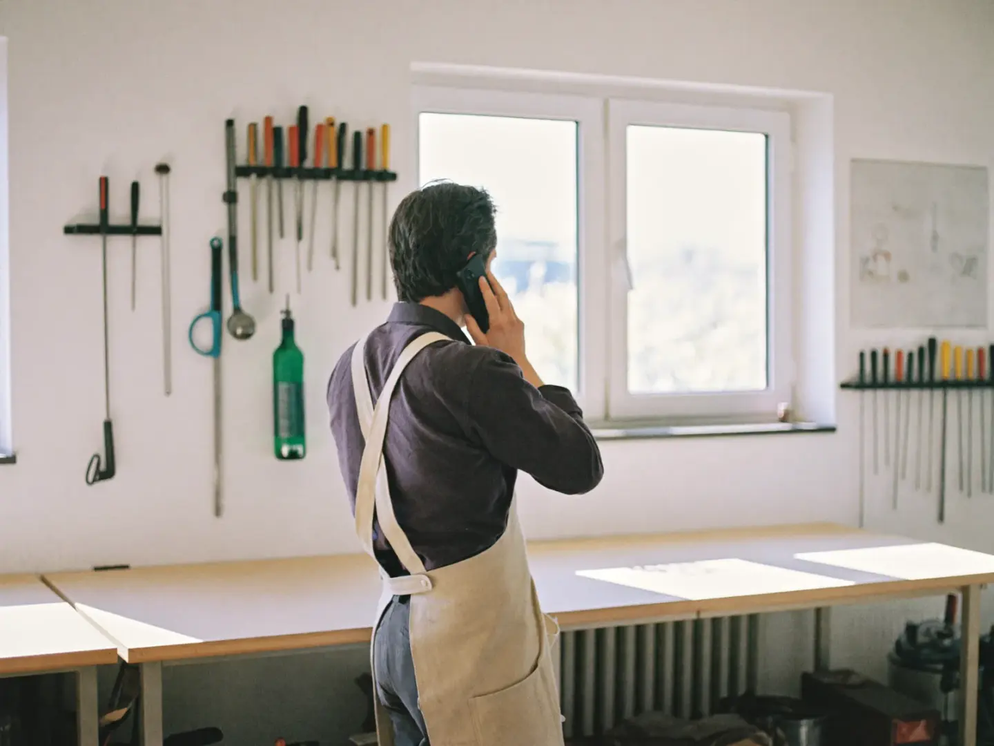 Handwerker in Arbeitsschürze telefoniert in seiner Werkstatt, an der Wand hängt geordnetes Werkzeug.