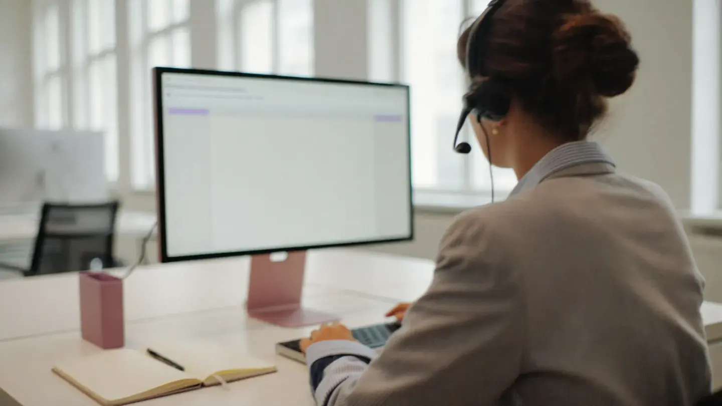 Mitarbeiterin mit Headset an einem hellen Schreibtisch, vor sich ein Monitor mit einer Vorgangsliste.