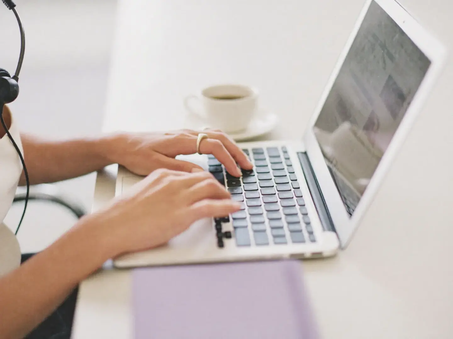 Hände tippen auf einer Laptop-Tastatur, daneben liegen ein Headset und eine Tasse Kaffee.